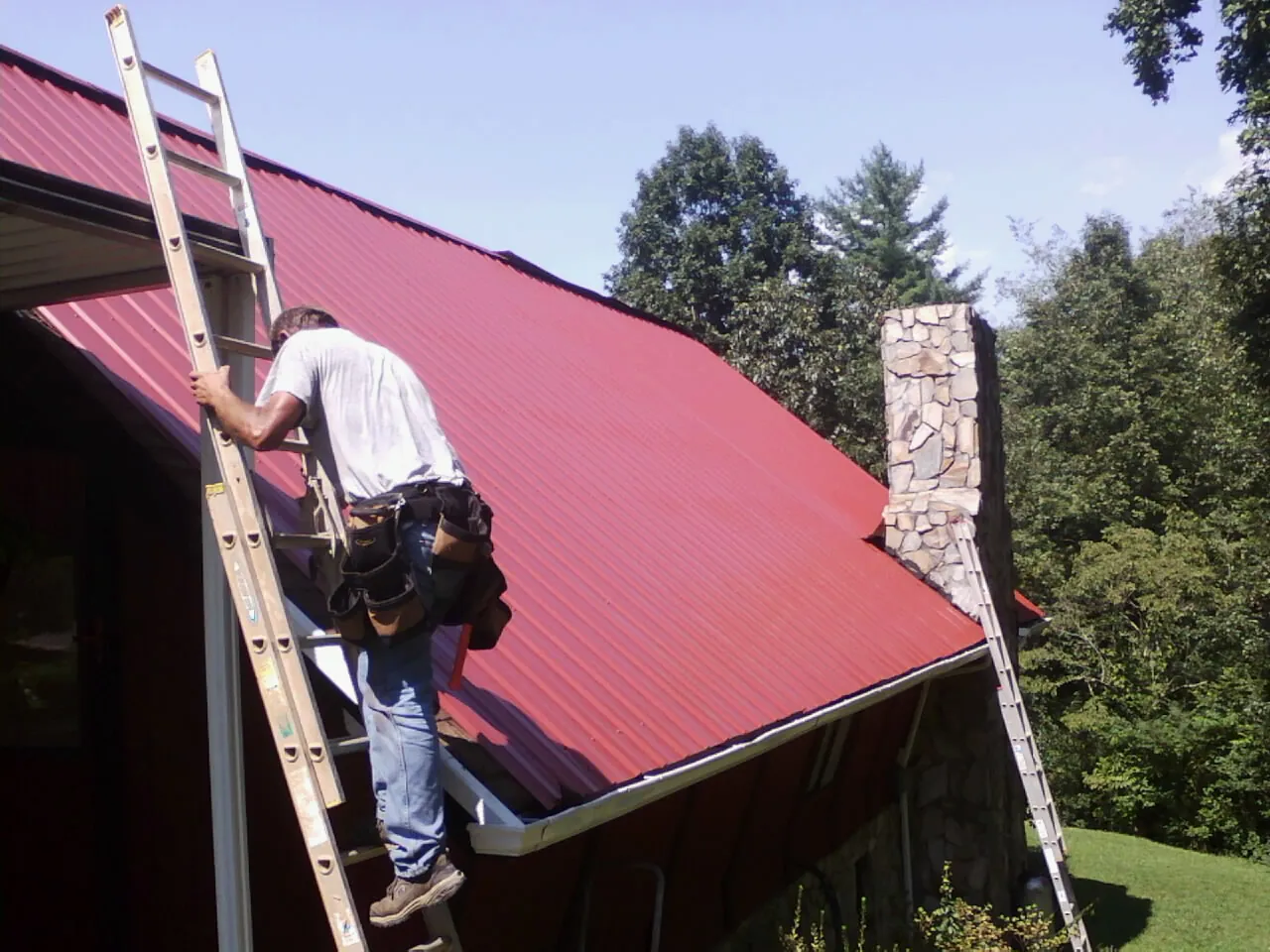 Roofer on a ladder beside a red metal roof and stone chimney — Soffit & Fascia Repair by Roofing Brothers in Grove